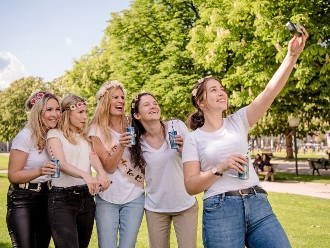 Fünf Frauen in weißen T-Shirts und Blumenkränzen machen ein Selfie im Freien. Sie halten Getränkedosen und lächeln in die Kamera., © ExitGames Stuttgart Fünf Frauen in weißen T-Shirts und Blumenkränzen machen ein Selfie im Freien. Sie halten Getränkedosen und lächeln in die Kamera., © ExitGames Stuttgart