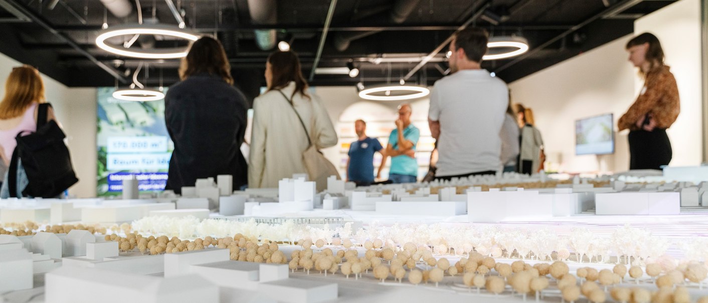 Besucher betrachten ein Architekturmodell in einer Ausstellung. Moderne Deckenleuchten und ein Bildschirm im Hintergrund., © Dominique Brewing Besucher betrachten ein Architekturmodell in einer Ausstellung. Moderne Deckenleuchten und ein Bildschirm im Hintergrund., © Dominique Brewing