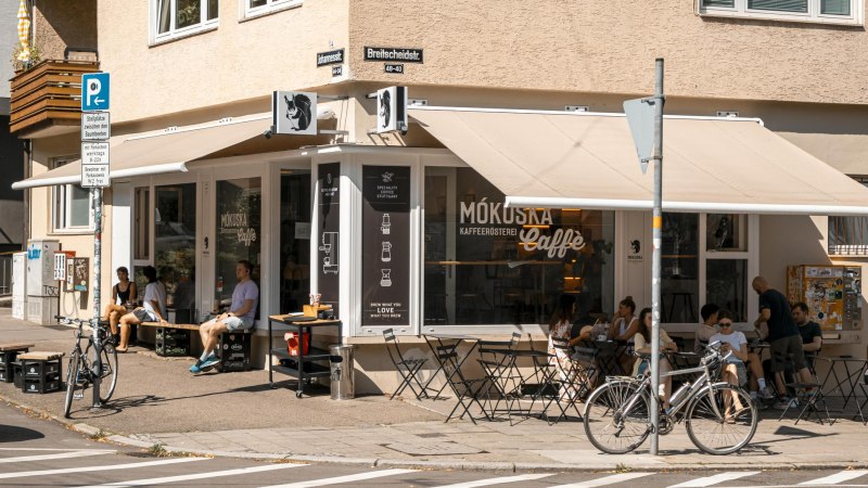 Café Mókuska an einer Straßenecke mit Außenbestuhlung und Gästen. Fahrräder sind geparkt, und es gibt Straßenschilder und einen Sonnenschutz., © SMG, Sarah Schmid