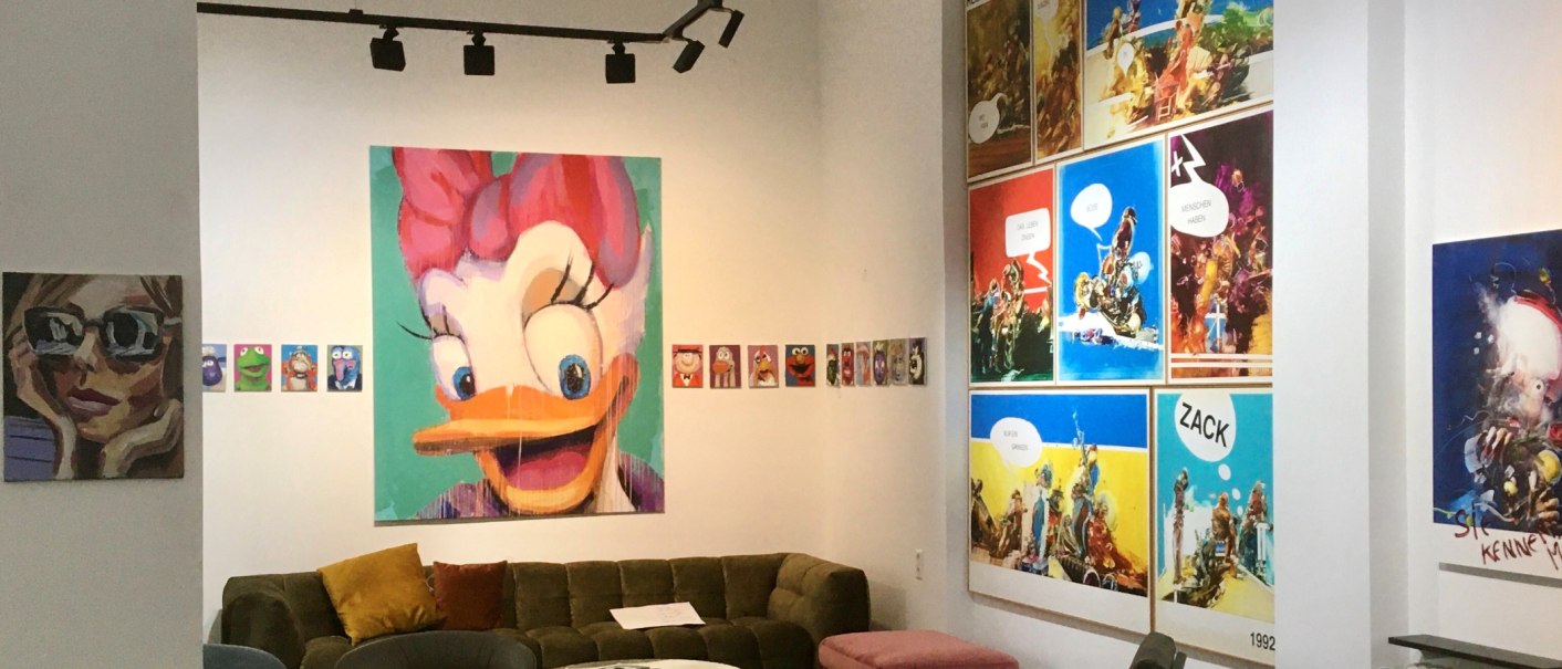 A gallery displays colorful Pop Art paintings of comic book characters and modern artworks. A large painting of a duck dominates the wall. There is a sofa in the room., © Galerie Z A gallery displays colorful Pop Art paintings of comic book characters and modern artworks. A large painting of a duck dominates the wall. There is a sofa in the room., © Galerie Z