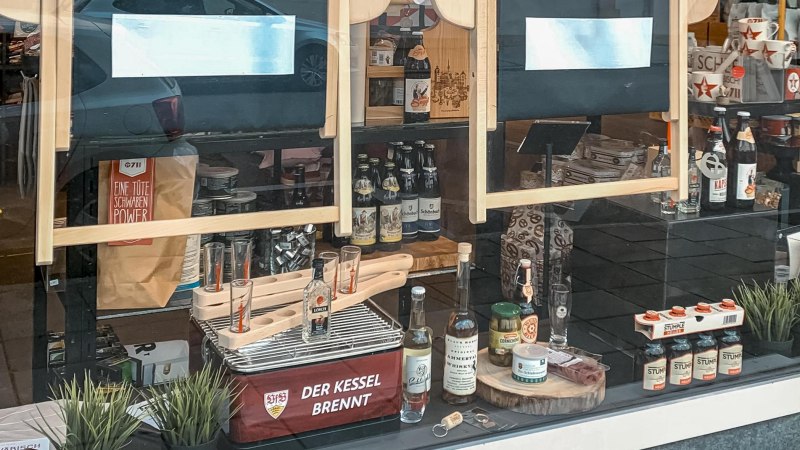 Schaufenster mit regionalen Produkten wie Bier, Spirituosen und Lebensmitteln. Dekoriert mit Pflanzen und Werbeschildern. Schriftzug 'Der Kessel brennt' sichtbar., © 0711 Store, Stuttgart