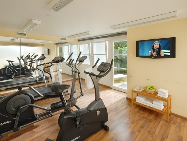 A bright fitness room with cardio equipment, a wall of mirrors, a TV on the wall and a shelf with towels and apples., &copy; relexa Waldhotel Schatten