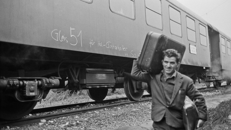 A man carries two suitcases next to a train with the inscription 'Gleis 51 für ital. Guest workers'., © Landesarchiv Baden-Württemberg HStAS Q 2/50 Nr. 2926-29