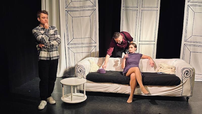 Eine Frau in grünem Kleid spricht auf einer Bühne, während ein Mann mit Hut neben ihr sitzt und zuschaut., © Theater Atelier Eine Frau in grünem Kleid spricht auf einer Bühne, während ein Mann mit Hut neben ihr sitzt und zuschaut., © Theater Atelier