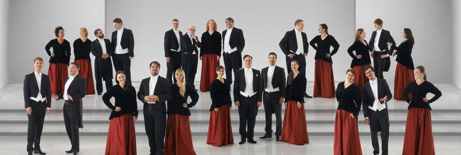 Der Kammerchor Stuttgart posiert in formeller Kleidung auf einer Bühne. Die Frauen tragen rote Röcke und schwarze Oberteile, die Männer Frack., © Jens Meisert