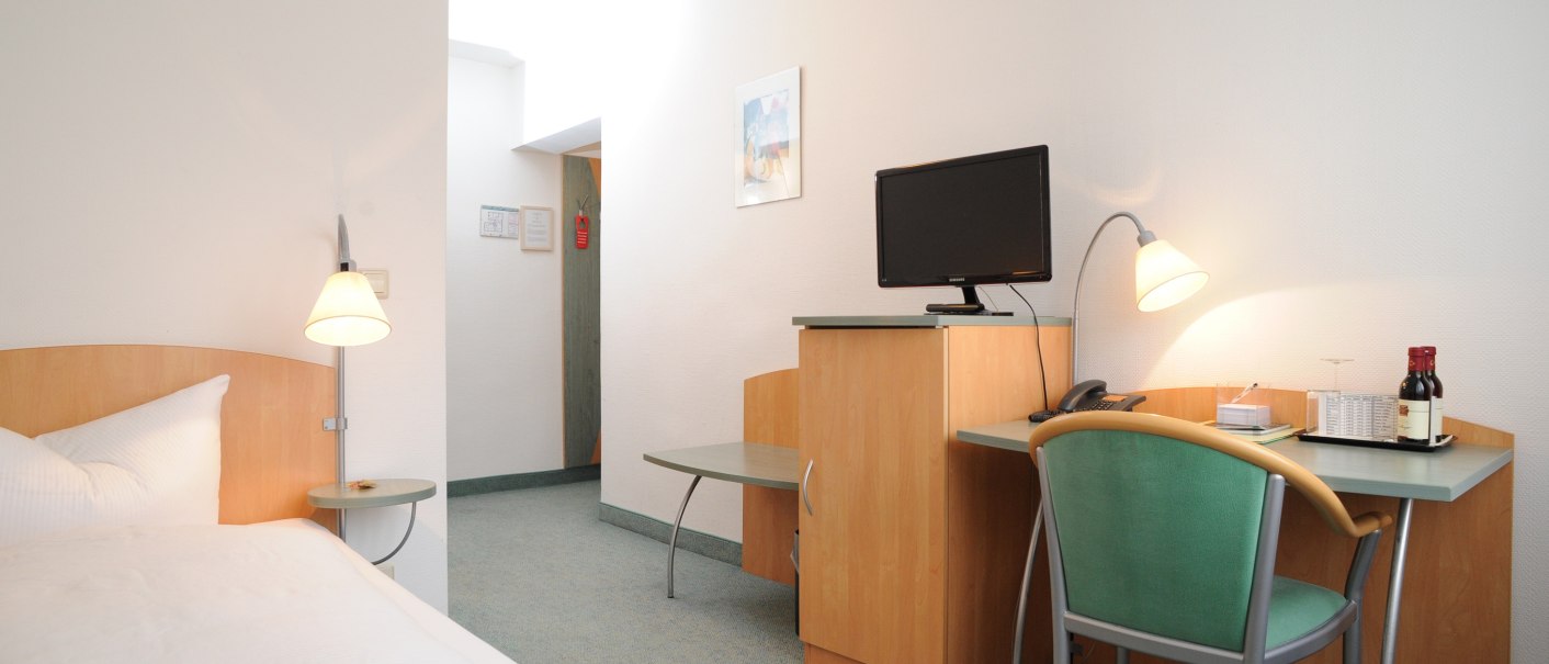 Bright single room with bed, desk, chair and TV. A lamp illuminates the desk, which also has drinks on it., © TOMAS