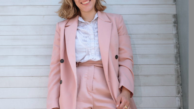 Eine lächelnde Person in einem rosa Anzug steht vor einer weißen Wand. Sie trägt ein weißes Hemd und hat blonde Haare., © Mafalda Rakoš Eine lächelnde Person in einem rosa Anzug steht vor einer weißen Wand. Sie trägt ein weißes Hemd und hat blonde Haare., © Mafalda Rakoš