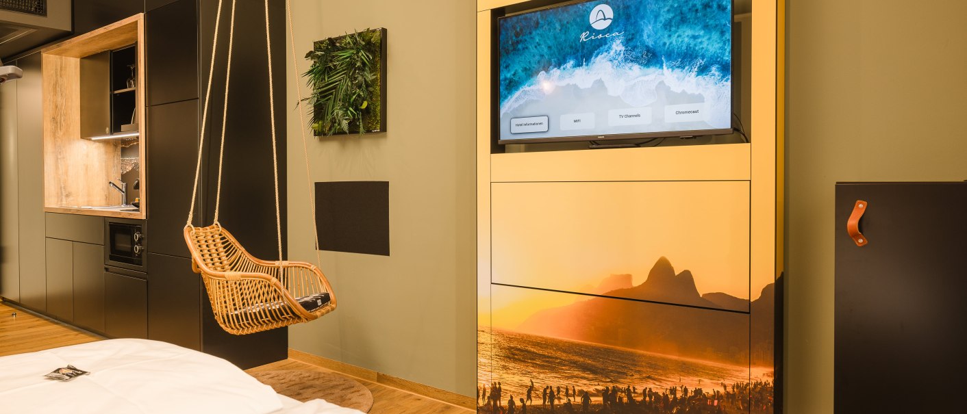 Modernes Hotelzimmer mit Hängesessel, Fernseher mit Strandbild, kleiner Küchenzeile und dekorativem Pflanzenbild an der Wand., © TOMAS