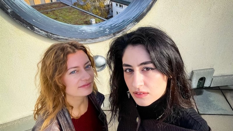 Zwei Frauen auf einem Dach vor einem runden Fenster, das den Blick auf eine Stadtlandschaft freigibt. Sie tragen lässige Kleidung und schauen in die Kamera., © Valerie Campbell Zwei Frauen auf einem Dach vor einem runden Fenster, das den Blick auf eine Stadtlandschaft freigibt. Sie tragen lässige Kleidung und schauen in die Kamera., © Valerie Campbell