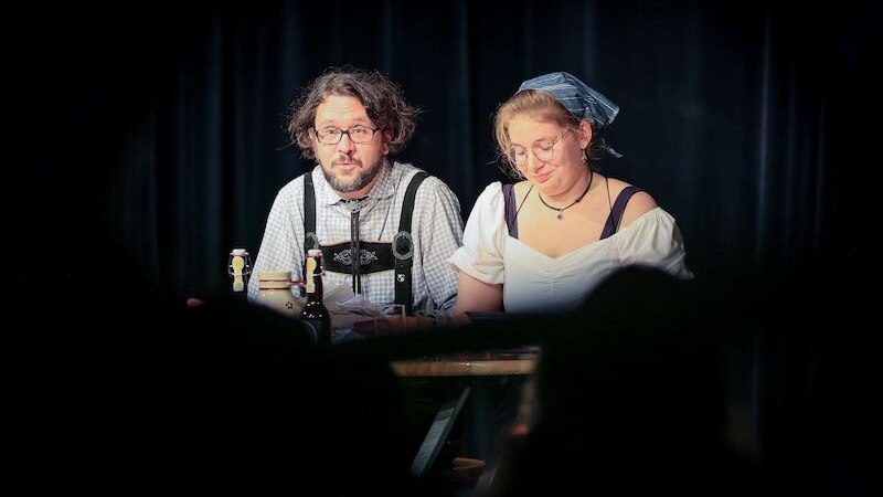 Zwei Personen in traditioneller Kleidung sitzen auf einer Bühne mit Mikrofonen. Der Mann trägt eine Brille und Hosenträger, die Frau ein Kopftuch., © Theaterhaus Stuttgart e.V. Zwei Personen in traditioneller Kleidung sitzen auf einer Bühne mit Mikrofonen. Der Mann trägt eine Brille und Hosenträger, die Frau ein Kopftuch., © Theaterhaus Stuttgart e.V.