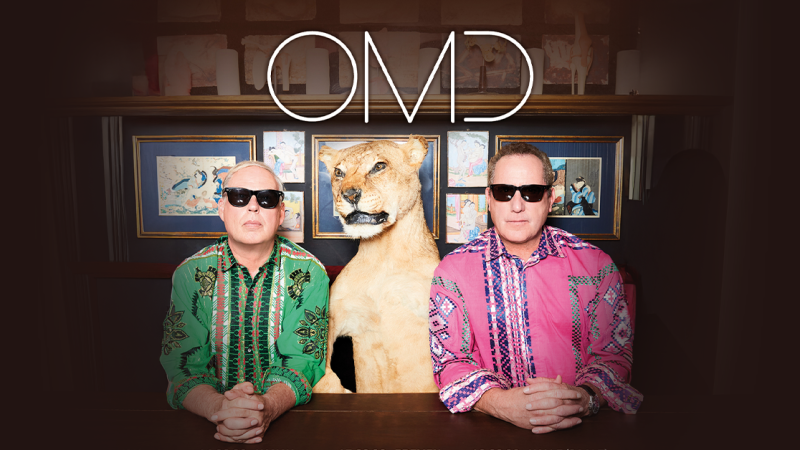 Zwei Männer in bunten Hemden und Sonnenbrillen sitzen neben einem ausgestopften Löwenkopf. Im Hintergrund sind gerahmte Bilder zu sehen. Oben steht 'OMD'., © links im Bild Zwei Männer in bunten Hemden und Sonnenbrillen sitzen neben einem ausgestopften Löwenkopf. Im Hintergrund sind gerahmte Bilder zu sehen. Oben steht 'OMD'., © links im Bild