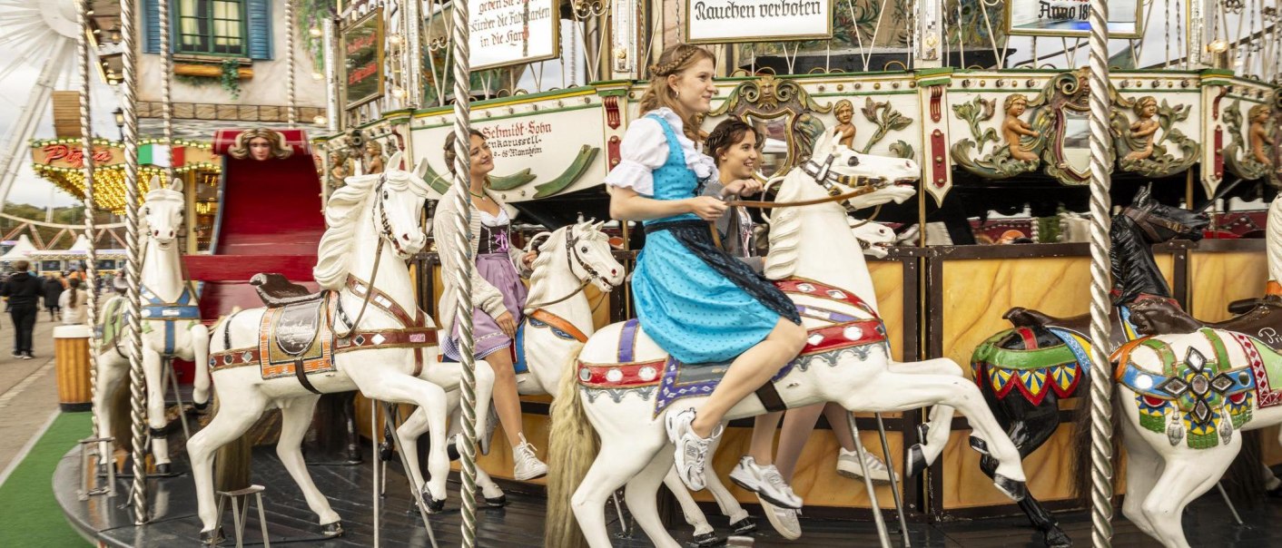 Two women in dirndls ride on a nostalgic carousel with white horses. A Ferris wheel and a pizza stand can be seen in the background., © Stuttgart-Marketing GmbH, Sarah Schmid Two women in dirndls ride on a nostalgic carousel with white horses. A Ferris wheel and a pizza stand can be seen in the background., © Stuttgart-Marketing GmbH, Sarah Schmid