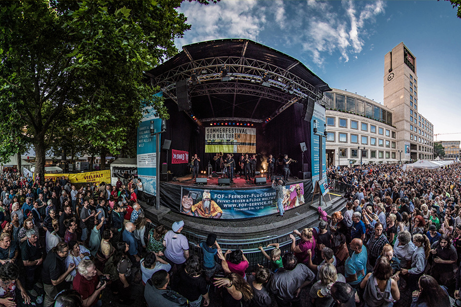 Eine große Menschenmenge genießt ein Konzert auf dem Sommerfestival der Kulturen. Die Bühne ist von Bäumen und Gebäuden umgeben., © Forum der Kulturen Stuttgart e. V. Eine große Menschenmenge genießt ein Konzert auf dem Sommerfestival der Kulturen. Die Bühne ist von Bäumen und Gebäuden umgeben., © Forum der Kulturen Stuttgart e. V.