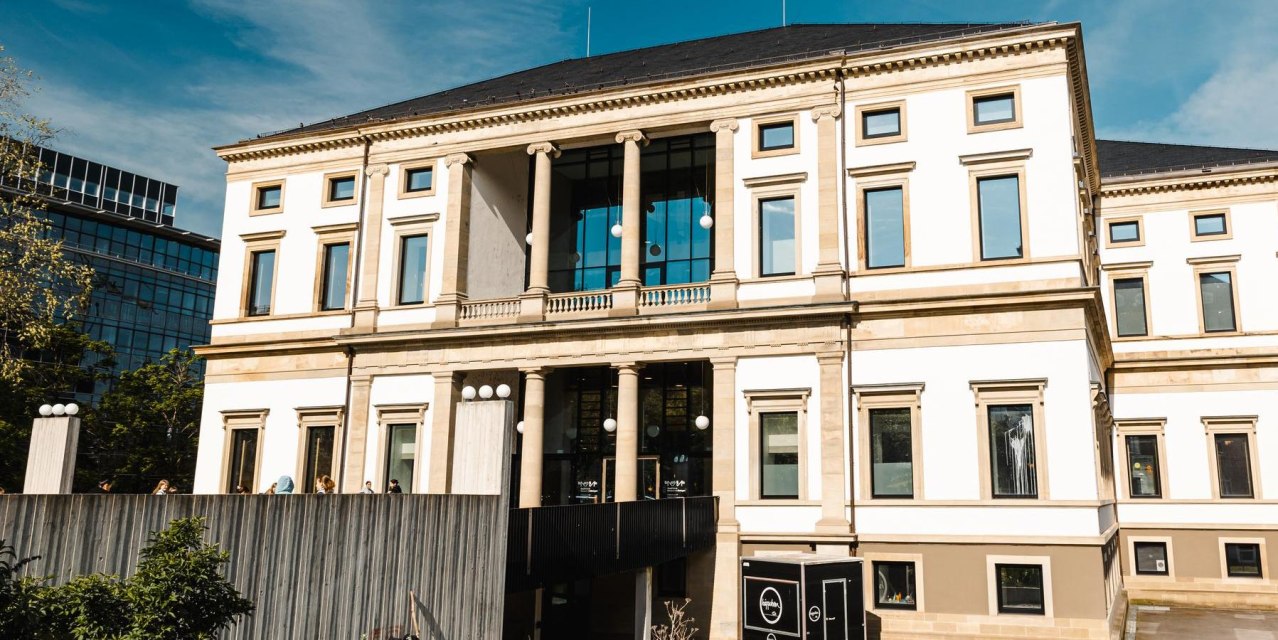 Ein historisches Gebäude mit Säulen und großen Fenstern, umgeben von moderner Architektur und Bäumen, unter einem klaren blauen Himmel., © Stuttgart Marketing GmbH, Sarah Schmid Ein historisches Gebäude mit Säulen und großen Fenstern, umgeben von moderner Architektur und Bäumen, unter einem klaren blauen Himmel., © Stuttgart Marketing GmbH, Sarah Schmid