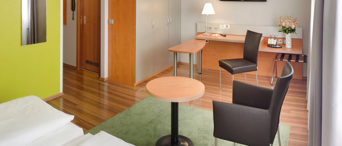 Modernes Hotelzimmer mit Doppelbett, Schreibtisch, Stühlen, Flachbildfernseher und grünem Teppich auf Holzboden., © attimo Hotel Stuttgart Modernes Hotelzimmer mit Doppelbett, Schreibtisch, Stühlen, Flachbildfernseher und grünem Teppich auf Holzboden., © attimo Hotel Stuttgart
