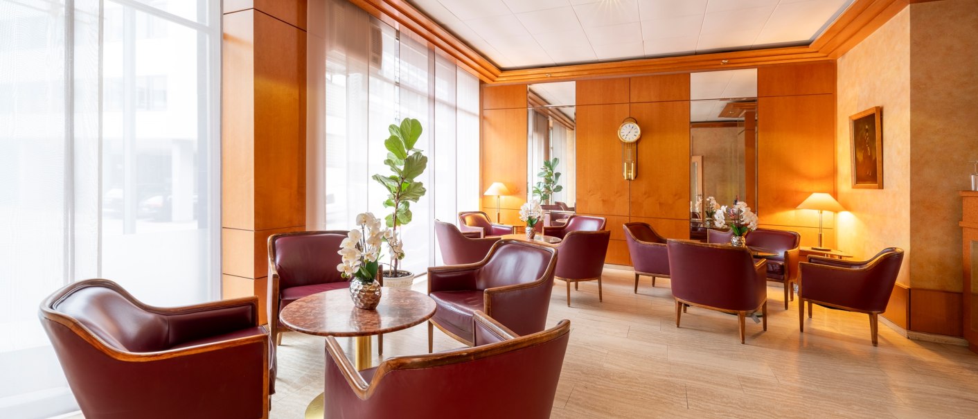 Elegant lounge with red leather armchairs, wooden walls and large windows. Plants and flowers on the tables, clock on the wall., © PLAZA INN Rieker Stuttgart Hauptbahnhof