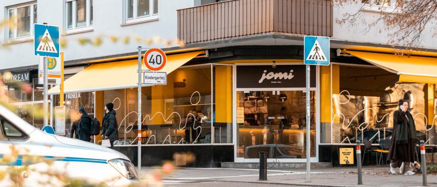 Schaufenster des Deli mit gelben Markisen. &Uuml;ber der T&uuml;r am Eck des Geb&auml;udes steht "jomi Bagel Deli"., &copy; Stuttgart-Marketing GmbH, Sarah Schmid