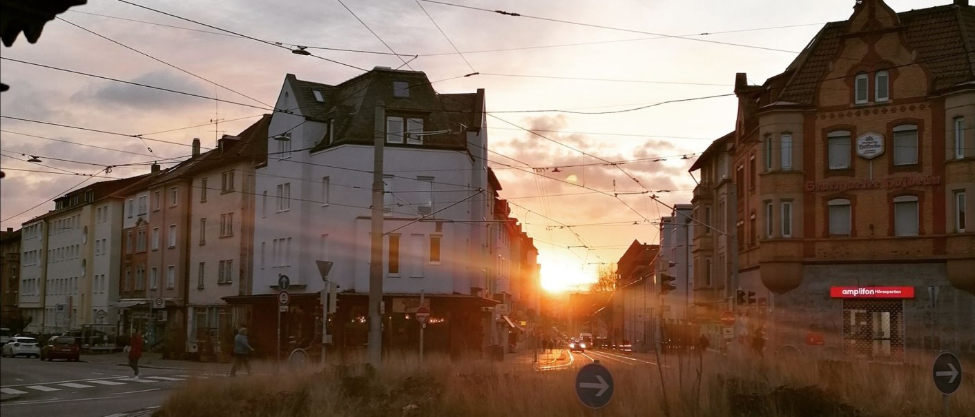 Sonnenuntergang in einer Stadtstraße mit historischen Gebäuden und Straßenbahnkabeln. Menschen überqueren die Straße, während die Sonne zwischen den Häusern untergeht., © SMG