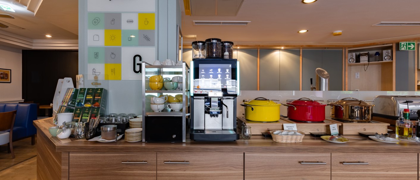 Ein Fr&uuml;hst&uuml;cksbuffet mit Kaffeemaschine, bunten T&ouml;pfen und Geschirr. Ein Schild mit der Aufschrift 'Make it yours.' ist sichtbar., &copy; Novum  Hospitality