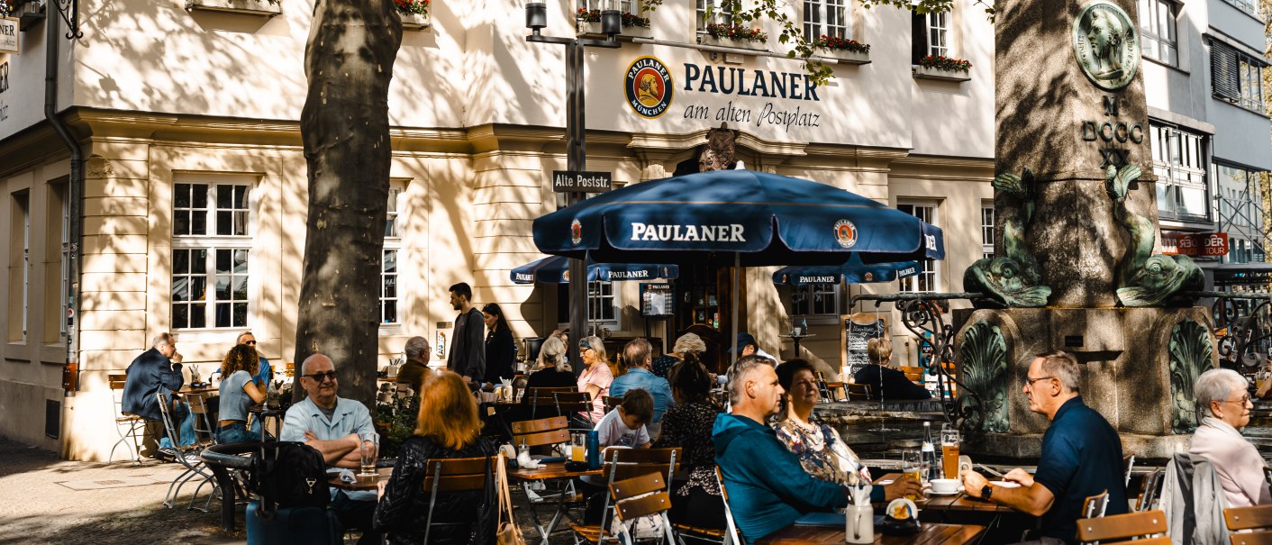 Menschen sitzen im Au&szlig;enbereich eines Paulaner-Restaurants. Ein gro&szlig;er Baum spendet Schatten, und ein Brunnen ist im Vordergrund zu sehen., &copy; SMG Stuttgart Marketing GmbH - Sarah Schmid