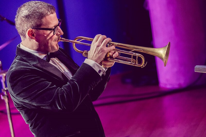Ein Mann in Anzug spielt Trompete auf einer Bühne mit lila Beleuchtung. Er trägt eine Brille und ist in eine musikalische Darbietung vertieft., © Württembergische Staatstheater Stuttgart