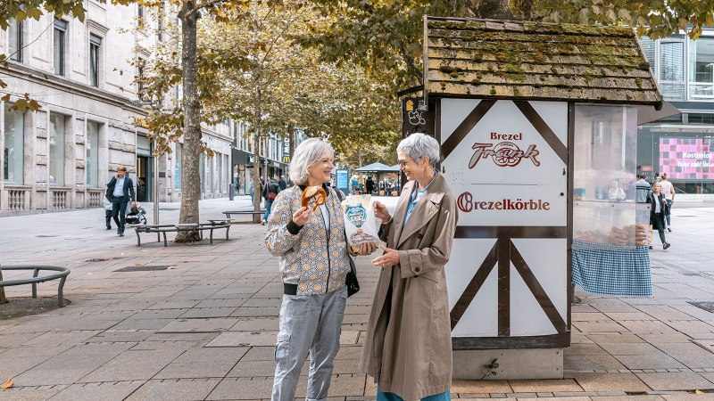 Zwei Frauen lachen vor einem Brezelstand am Schlossplatz. Eine hält eine Brezel, die andere eine Tüte. Herbstliche Bäume im Hintergrund., © SMG, Sarah Schmid