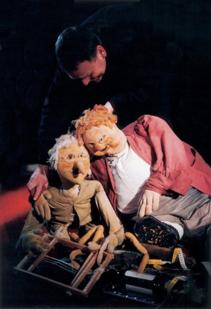 Ein Puppenspieler manipuliert zwei große Puppen, die auf dem Boden sitzen. Eine Puppe trägt eine rote Jacke, die andere eine beige Kleidung., © FITZ Das Theater animierter Formen