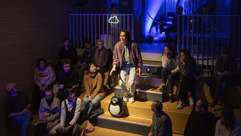 Ein Mann steht in einem kleinen Theater und spricht zu einer Gruppe von Menschen. Die Bühne ist blau beleuchtet., © tri-bühne e.V.