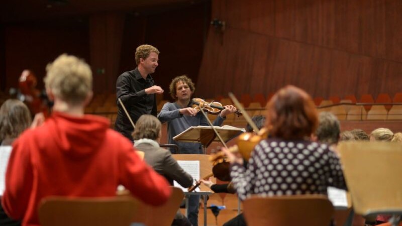 Ein Dirigent und ein Violinist leiten eine Orchesterprobe in einem Konzertsaal. Musiker sitzen auf St&uuml;hlen und spielen Instrumente., &copy; il Gusto Barocco - Stuttgarter Barockorchester