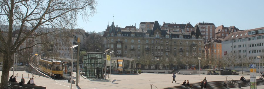 Marienplatz in Stuttgart mit Straßenbahn, modernen und historischen Gebäuden. Menschen sitzen auf Treppen, Bäume ohne Laub, sonniger Tag., © Stuttgart-Marketing GmbH