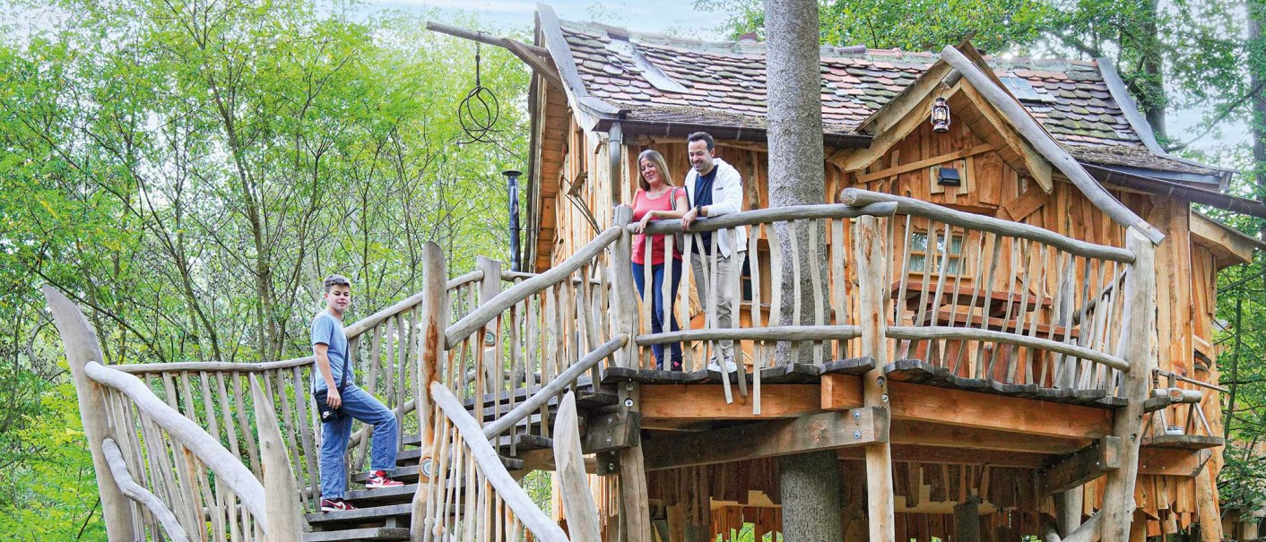 A rustic treehouse with a wooden railing. Three people are standing on the porch, surrounded by green trees., &copy; Tripsdrill Natur-Resort GmbH