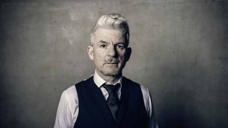 A man with gray hair wears a suit and stands in front of a gray wall. He looks seriously into the camera., &copy; Theaterhaus Stuttgart e.V.