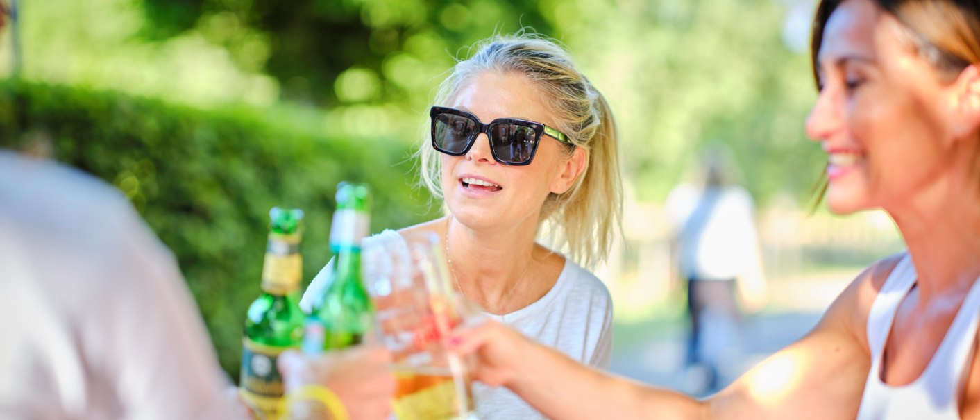 Three people are toasting with drinks outdoors. One woman is wearing sunglasses and smiling. The scene has a relaxed, summery feel., © Katzenbacher Hof Three people are toasting with drinks outdoors. One woman is wearing sunglasses and smiling. The scene has a relaxed, summery feel., © Katzenbacher Hof