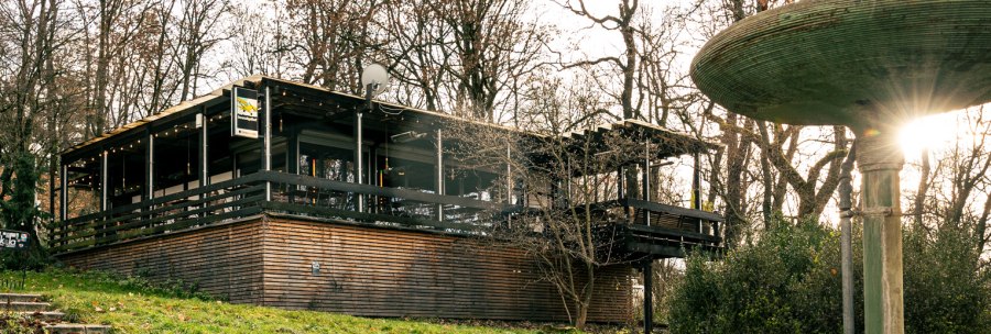 Building with wooden cladding and terrace in wooded surroundings, next to a large, multi-tiered fountain. Rays of sunlight shine through the trees., © Stuttgart Marketing GmbH © Sarah Schmid Building with wooden cladding and terrace in wooded surroundings, next to a large, multi-tiered fountain. Rays of sunlight shine through the trees., © Stuttgart Marketing GmbH © Sarah Schmid