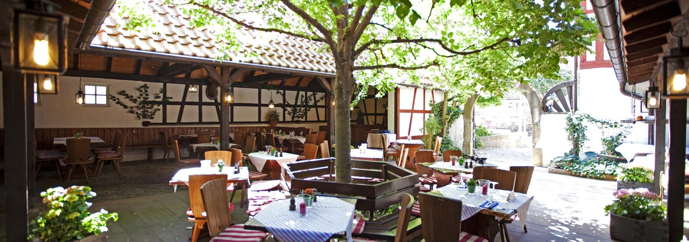 Ein gemütlicher Innenhof mit Tischen und Stühlen unter einem Baum, umgeben von Fachwerkhäusern. Die Tische sind mit gestreiften Tischdecken gedeckt., © Hotel Hirsch