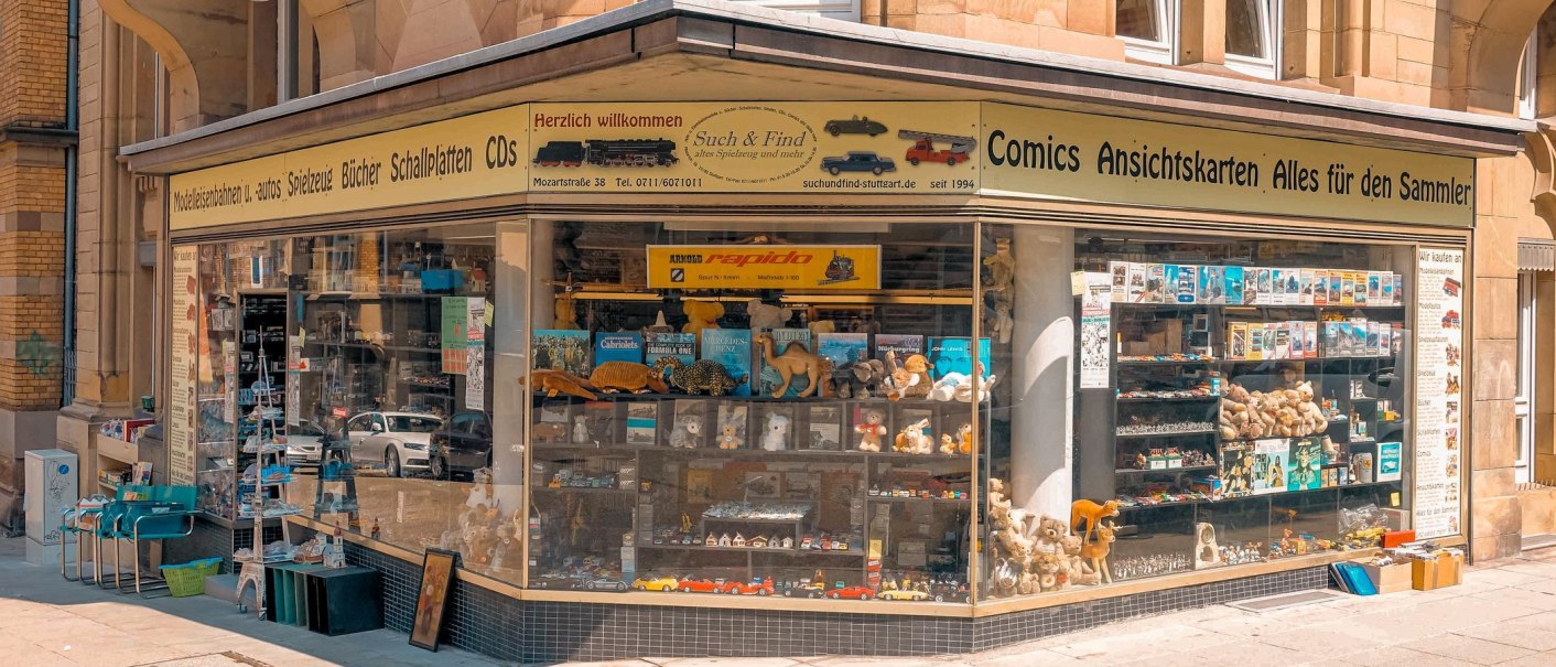Eckgeschäft mit Schaufenstern voller Spielzeug, Modellautos und Büchern. Oben steht 'Such & Find' und 'Comics Ansichtskarten Alles für den Sammler'., © Such & Find, Stuttgart