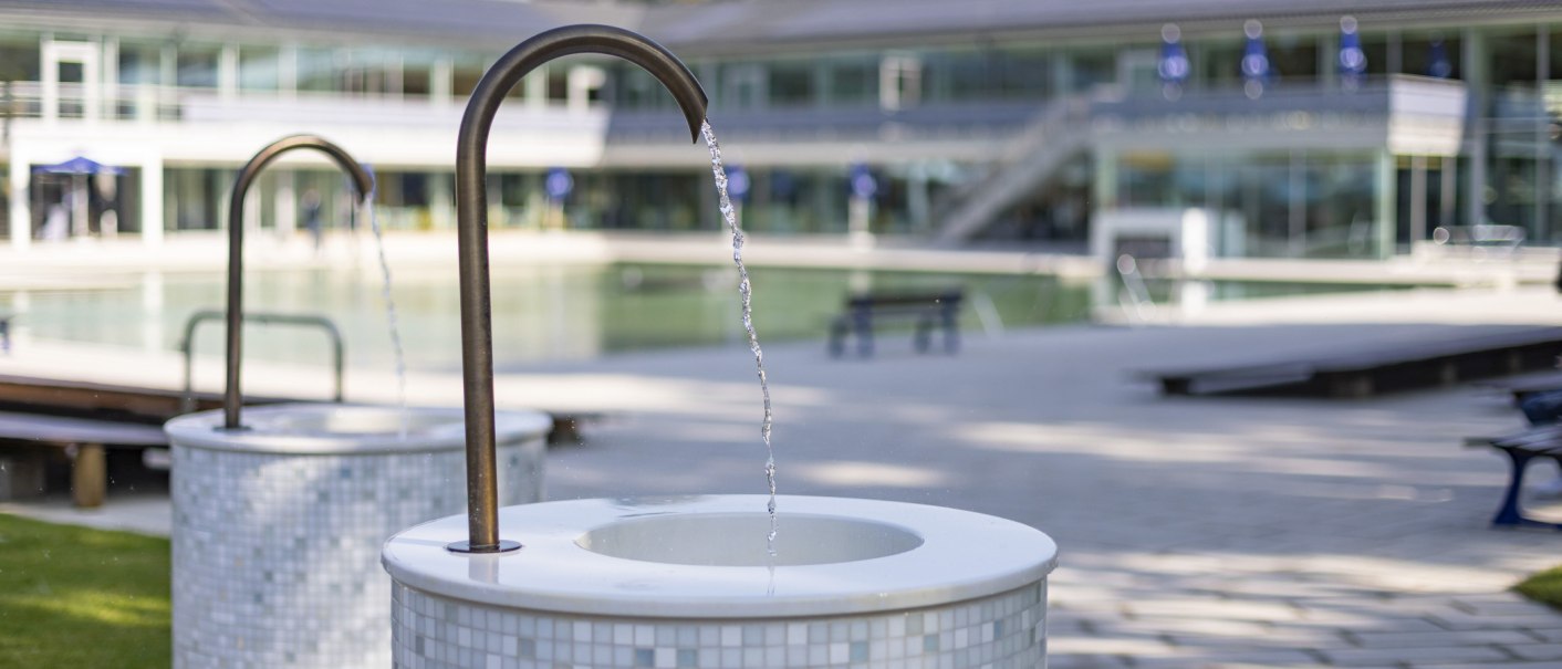 Ein Trinkbrunnen im Mineralbad Berg mit flie&szlig;endem Wasser im Vordergrund, im Hintergrund ein modernes Geb&auml;ude und ein Pool., &copy; Stuttgarter B&auml;der