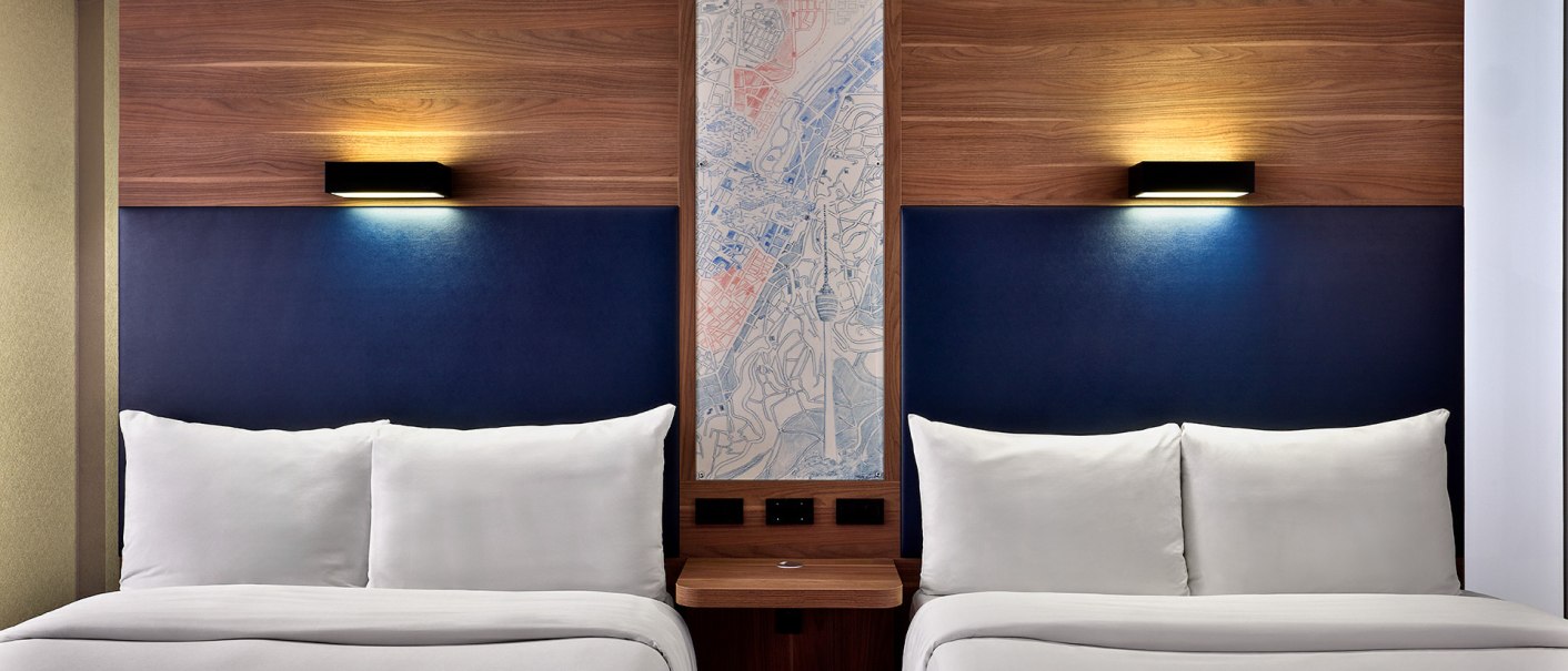 Two hotel beds with blue upholstery and wooden headboard, separated by a narrow table. A map is attached to the wall between the beds., © Aloft Stuttgart Two hotel beds with blue upholstery and wooden headboard, separated by a narrow table. A map is attached to the wall between the beds., © Aloft Stuttgart
