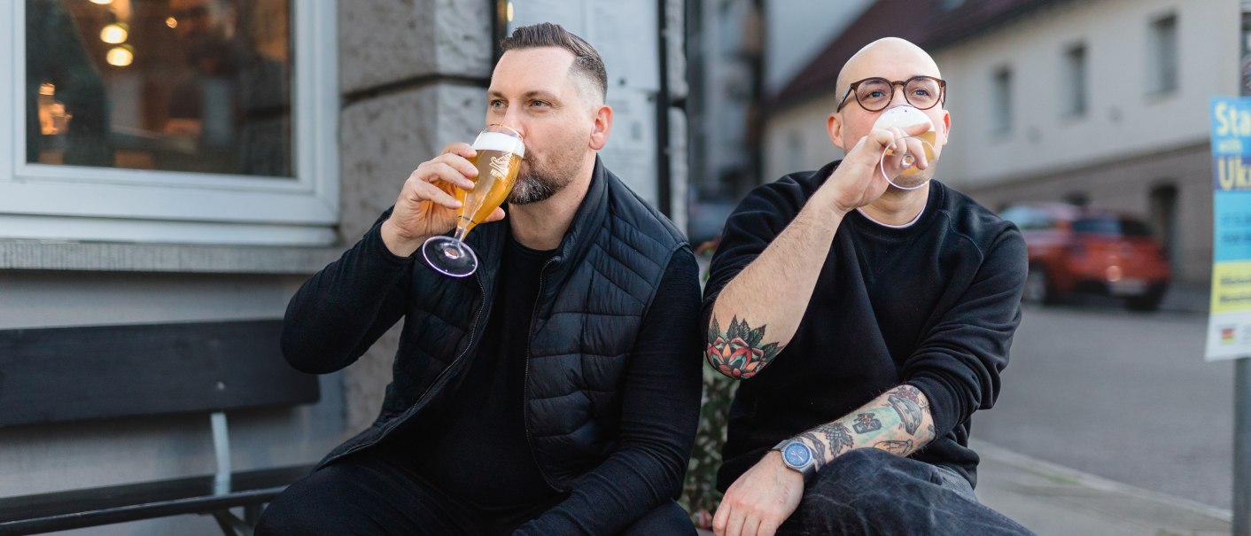 Two men are sitting on a wooden pallet in front of a building drinking beer. One of them wears glasses and has tattooed arms., © © Jaimee Moses All Rights Reserved Two men are sitting on a wooden pallet in front of a building drinking beer. One of them wears glasses and has tattooed arms., © © Jaimee Moses All Rights Reserved