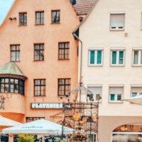 The Hans im Glück fountain in front of historic buildings. Parasols and figures can be seen in the foreground. The houses have red roofs and many windows., © SMG, takemyhearteverywhere The Hans im Glück fountain in front of historic buildings. Parasols and figures can be seen in the foreground. The houses have red roofs and many windows., © SMG, takemyhearteverywhere