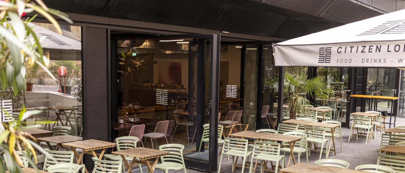 Outdoor area of a café with wooden tables and green chairs. An awning bears the lettering 'Citizen Long'. Plants decorate the area., © SMG, Sarah Schmid
