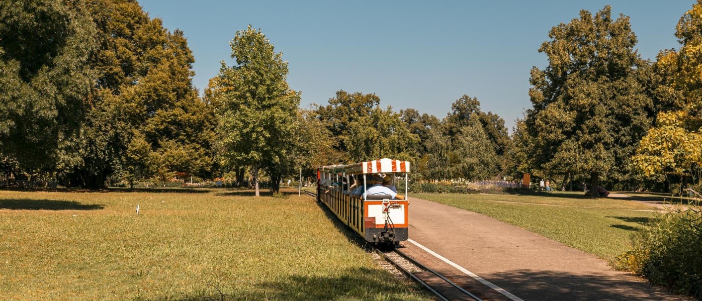 Eine kleine Bahn fährt durch einen grünen Park mit Bäumen und Wiese unter klarem Himmel., © Stuttgart-Marketing GmbH, Sarah Schmid