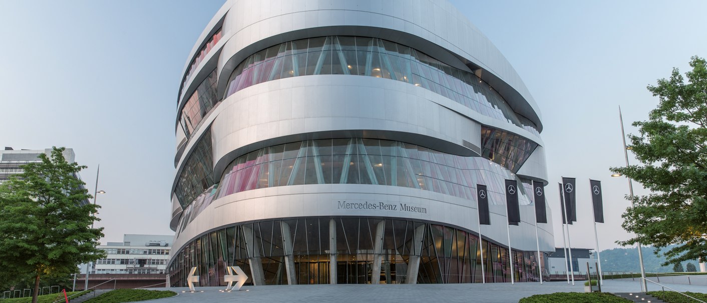 Das Mercedes-Benz Museum mit seiner modernen, geschwungenen Architektur und mehreren Mercedes-Flaggen davor., © Daimler AG Das Mercedes-Benz Museum mit seiner modernen, geschwungenen Architektur und mehreren Mercedes-Flaggen davor., © Daimler AG