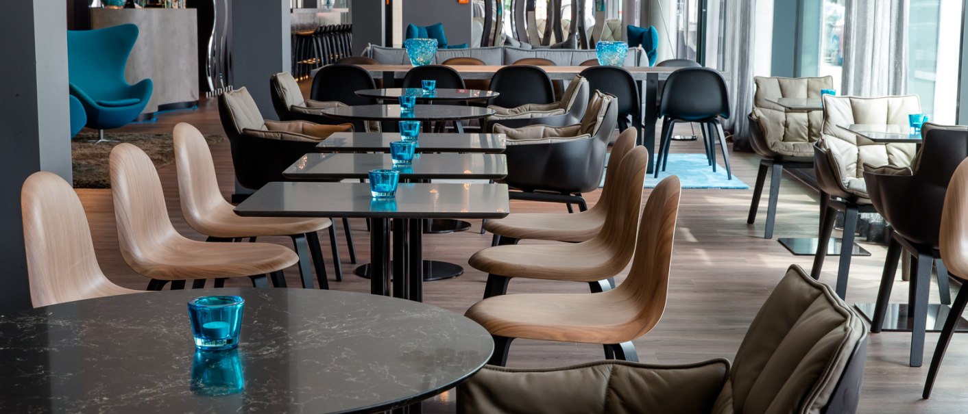 Moderner Fr&uuml;hst&uuml;cksbereich mit eleganten Tischen, St&uuml;hlen und blauen Akzenten. Stilvolle Beleuchtung und gro&szlig;e Fenster sorgen f&uuml;r eine einladende Atmosph&auml;re., &copy; Motel One Stuttgart-Bad Cannstatt