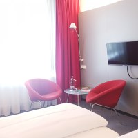 Hotelzimmer mit Doppelbett, roten Sesseln, kleinem Tisch, Wand-TV und Schreibtisch mit Kaffeemaschine. Helle Vorhänge und rote Akzente., © Vienna House Easy Mo. Stuttgart