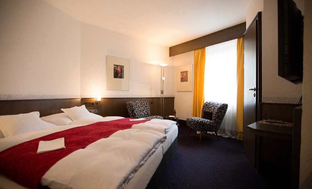 A cozy hotel room with a double bed, red accents, two armchairs, yellow curtains, and a wall-mounted TV., &copy; Hotel B&uuml;rkle