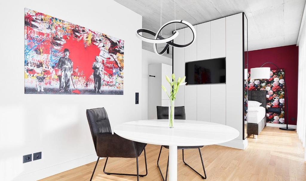 Moderner Essbereich mit rundem Tisch, zwei schwarzen Stühlen, auffälligem Kunstwerk an der Wand und Blick ins Schlafzimmer mit Blumenmuster., © livisit bergapartments Moderner Essbereich mit rundem Tisch, zwei schwarzen Stühlen, auffälligem Kunstwerk an der Wand und Blick ins Schlafzimmer mit Blumenmuster., © livisit bergapartments