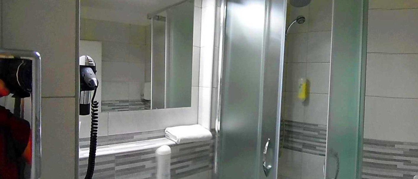 Modernes Badezimmer mit Dusche, Spiegel, Haartrockner und Handtuch. Die Wände sind mit grauen Fliesen dekoriert., © Hotel Arcis