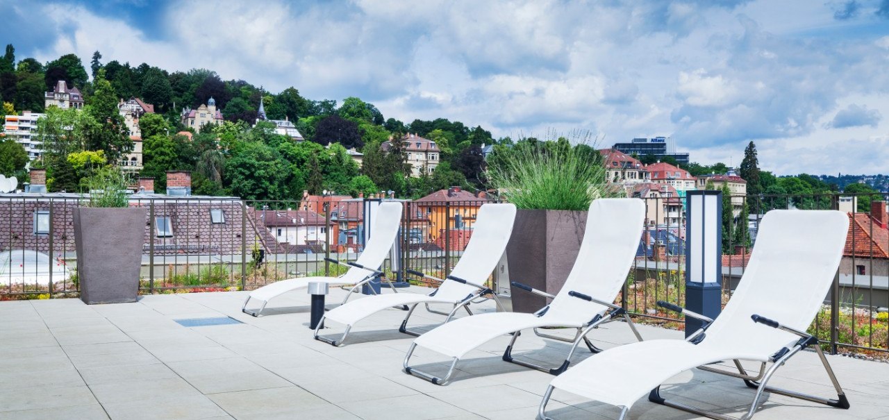 Dachterrasse mit weißen Liegestühlen, Pflanzen und Blick auf grüne Hügel und Häuser im Hintergrund., © Park Inn by Radisson Stuttgart