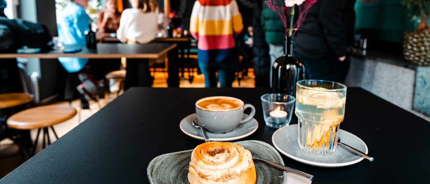 Ein Tisch in einem Café mit Zimtschnecke, Cappuccino und Zitronenwasser. Im Hintergrund sind unscharfe Personen zu sehen., © Stuttgart-Marketing GmbH, Sarah Schmid